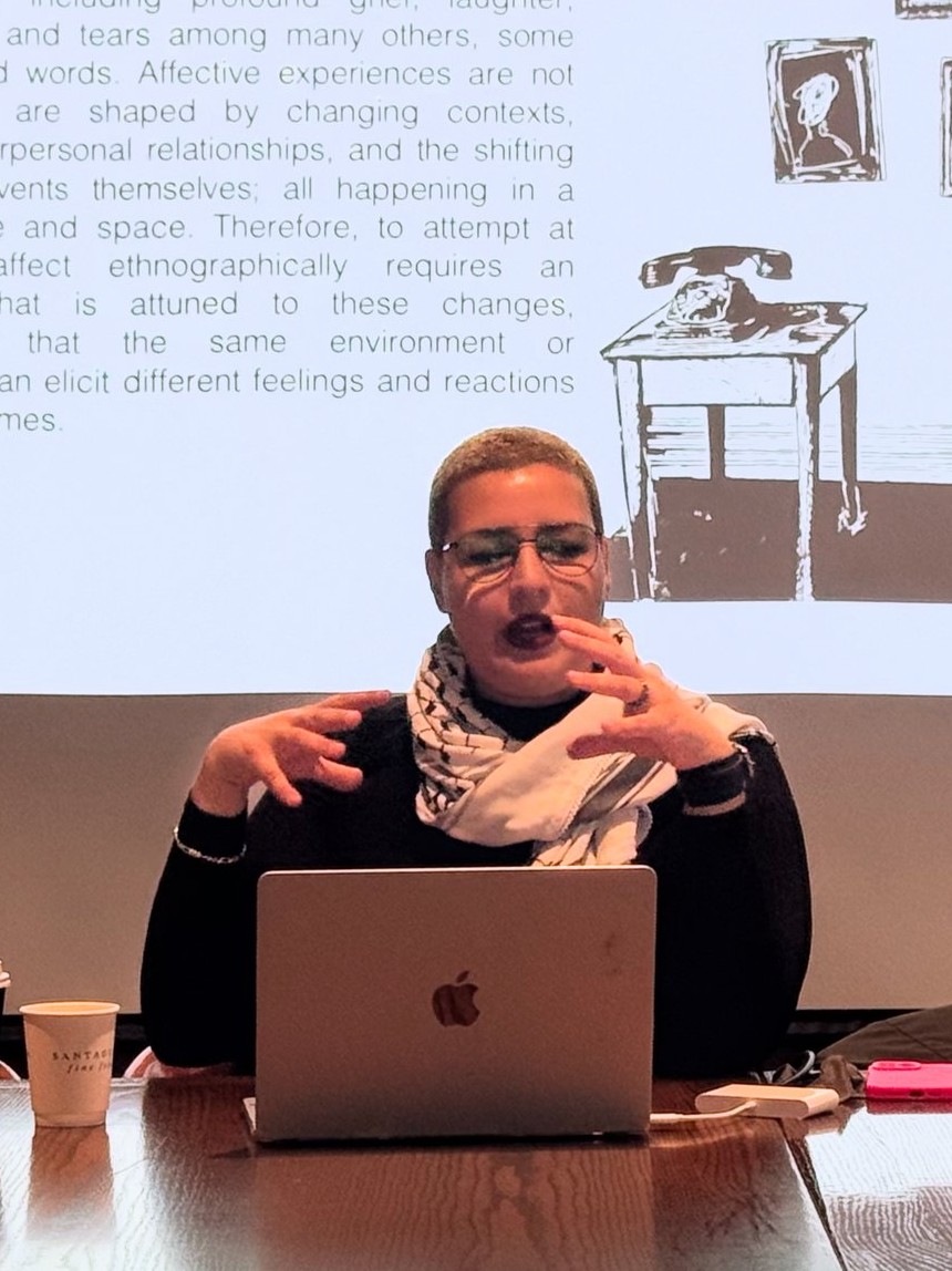 Maya El Helou seated at a table with a laptop, presenting to an audience beside a projected slide on affect and ethnographic fieldwork
