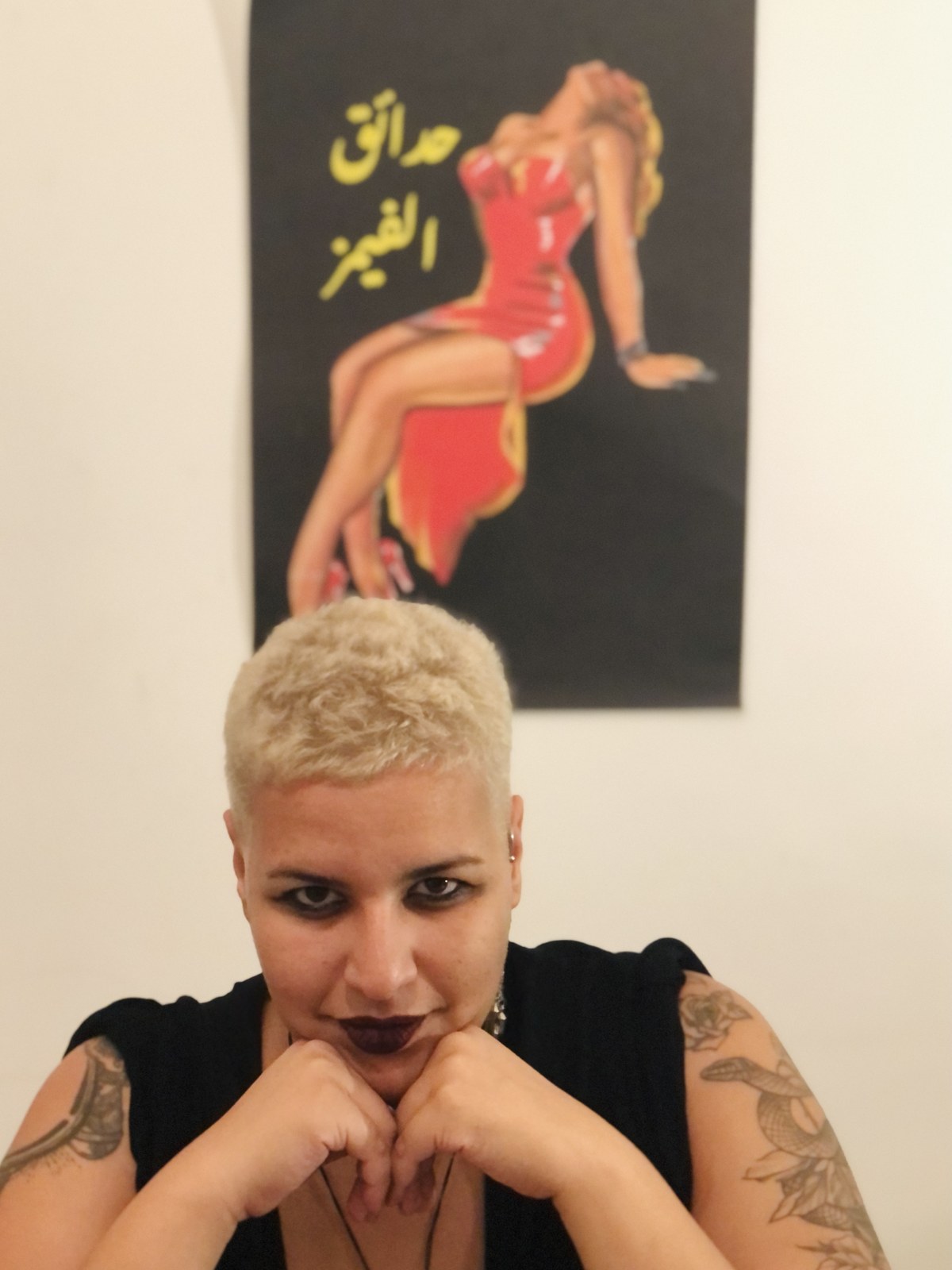 Maya El Helou seated with chin resting on their hands, in front of one of their posters