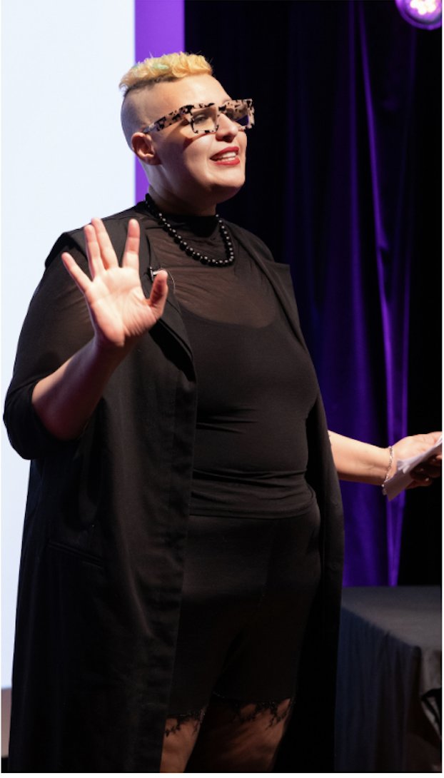 Maya El Helou standing and waving at an event, dressed in black with a beaded necklace
